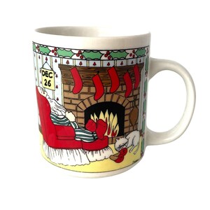 May include: A white ceramic mug featuring a colorful illustration of a person in a red armchair by a fireplace with red stockings hanging above. A calendar on the wall reads DEC 26. A white cat sleeps on the floor near a pair of red slippers.