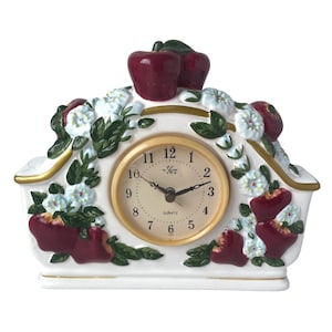 May include: A white ceramic mantel clock featuring a central round clock face with black numerals and hands. The clock is decorated with raised, hand-painted red apples, green leaves, and white blossoms, accented with gold trim along the edges.