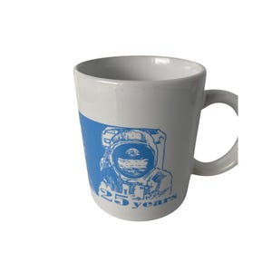 May include: White ceramic mug with a blue graphic of an astronaut and the text "25 years". The mug has a curved handle and a cylindrical shape. The design is on a blue background.
