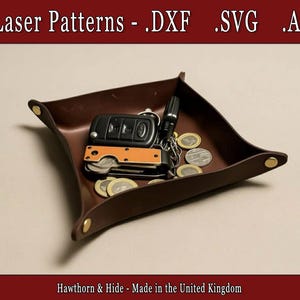 May include: A brown leather catchall tray with gold rivets at each corner. The tray contains car keys, a small knife, coins, and a flashlight. The text "Laser Patterns - .DXF .SVG .Ai" is at the top. The text "Hawthorn & Hide - Made in the United Kingdom" is at the bottom.