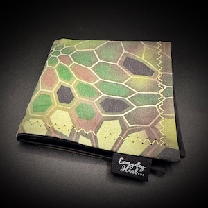 Puede incluir: Un pañuelo plegado con un diseño de camuflaje en tonos verdes, marrones y negros. Los bordes están cosidos con hilo amarillo. Una pequeña etiqueta negra dice "Everyday Hank EDC".