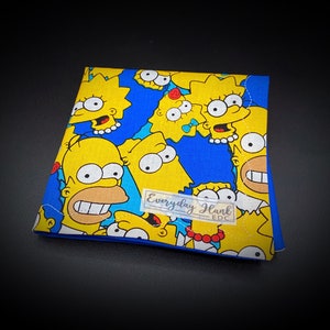 Puede incluir: Un cuadrado de tela azul con un patrón repetido de personajes de dibujos animados amarillos. Los personajes son de la serie de televisión Los Simpson. La tela tiene un borde cosido blanco. El texto "Everyday Hank EDC" está impreso en una pegatina transparente en la tela.