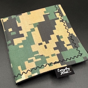 May include: A square fabric handkerchief featuring a digital camouflage pattern in shades of olive green, tan, and black. The edges are finished with a black zigzag stitch, and a small black fabric tag displays the white text Everyday Hank EDC.
