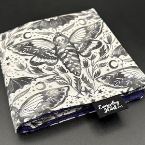 Puede incluir: Una bolsa de tela blanca con un estampado en blanco y negro de polillas, murciélagos y estrellas. La bolsa está doblada y tiene una etiqueta negra que dice "Everyday Hank EDC".