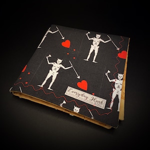 Puede incluir: Cartera de tela negra con un corazón rojo y un diseño de esqueleto. La cartera tiene un ribete de cuero marrón y el texto "Everyday Hank EDC" impreso en la parte delantera.