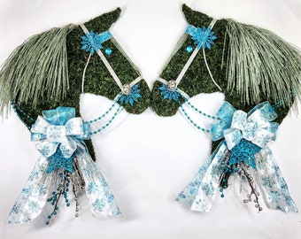 May include: Two horse head-shaped wreaths. The wreaths are covered in green material, with silver and blue accents. Each horse head has a silver bridle, blue snowflake decorations, and a large bow with snowflake patterns. The manes are made of silver tinsel.