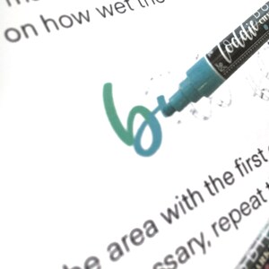 May include: A turquoise blue marker with a black cap and the word "loddit" written on the side. The marker is being used to write the letter "b" in green and turquoise blue on a white piece of paper.