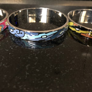 Op de afbeelding: Drie zilveren armbanden met kleurrijke emaille ontwerpen. Eén armband heeft een rood, roze en groen bloemenpatroon. Een andere heeft een blauw en lichtblauw golfontwerp. De derde armband heeft een geel, oranje en zwart ontwerp.