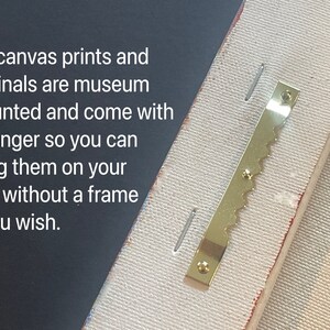 May include: Close-up of a gold-colored sawtooth hanger attached to the back of a canvas print. The hanger is designed to hang the print on a wall without a frame.