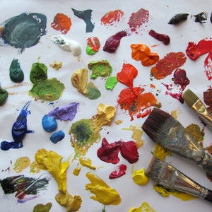 May include: A white surface with various colors of paint, including green, blue, red, yellow, and orange. Three paintbrushes are on the surface, two with brown handles and one with a silver handle.