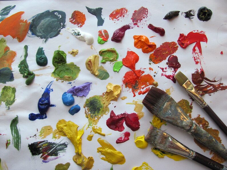 May include: A white surface with various colors of paint, including green, blue, red, yellow, and orange. Three paintbrushes are on the surface, two with brown handles and one with a silver handle.