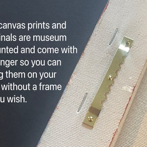 May include: Close-up of a gold-colored sawtooth hanger attached to the back of a canvas print. The hanger is designed to hang the print on a wall without a frame.