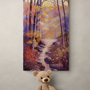 May include: A crocheted teddy bear with brown fur and cream-colored paws sits on a wooden table with fall leaves and chestnuts. A painting of a forest path with a stream and rocks hangs on the wall behind the bear.