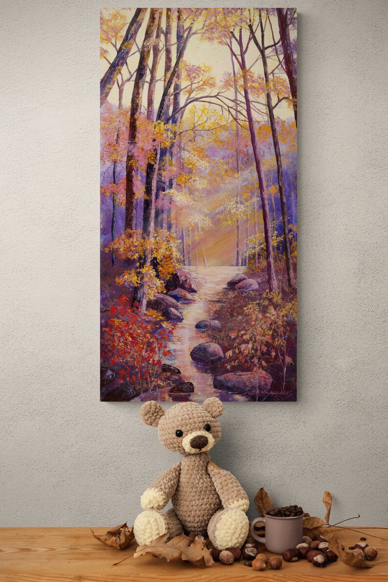 May include: A crocheted teddy bear with brown fur and cream-colored paws sits on a wooden table with fall leaves and chestnuts. A painting of a forest path with a stream and rocks hangs on the wall behind the bear.