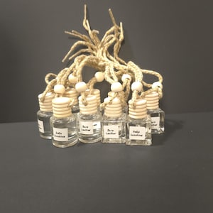 May include: A collection of small, clear glass bottles with wooden caps and labels. The labels read "Hello Sunshine" and "Bare Leather." The bottles are connected by a natural fiber string with wooden beads, suggesting a fragrance or aromatherapy product.