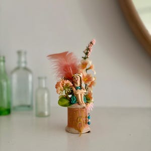 Op de afbeelding: Decoratief beeldje van een zittende fee bovenop een houten spoel, versierd met bloemen en een perzikkleurige veer. De fee draagt een turquoise jurk en een bloemenkroon. Een kralenketting hangt aan de spoel. Het beeldje staat tegen een vage achtergrond.