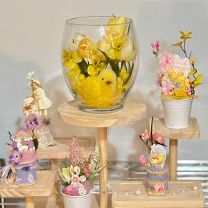May include: A wooden display stand features Easter-themed decorations. A large glass bowl holds yellow tulips, pansies, and a yellow chick. Other decorations include miniature buckets with flowers, eggs, and a bunny. The display is set against a neutral background.