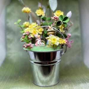 Può includere: Un piccolo secchio argentato lucido pieno di fiori gialli, verde e figurine in miniatura. La composizione include un quadrifoglio e una piccola statuina di coniglio. Lo sfondo è verde tenue.