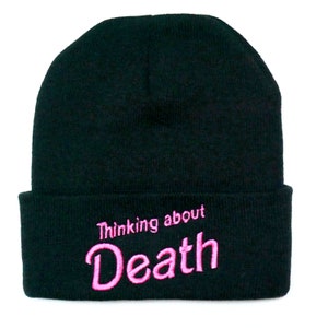 May include: Black beanie hat with the embroidered text "Thinking about Death" in pink. The hat is a classic knit style, suitable for cold weather. The text is in a playful, cursive font.