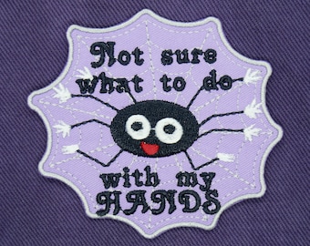 Op de afbeelding: Paarse stof met een geborduurde patch in de vorm van een lichtpaars spinnenweb. De spin heeft zwarte ogen en een rode mond. De tekst op de patch luidt "Not sure what to do with my HANDS".