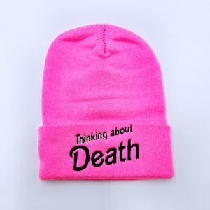 May include: A bright pink beanie with the text "Thinking about Death" embroidered in black.