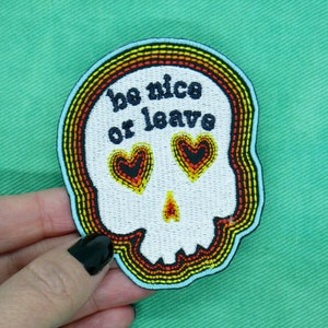 May include: Embroidered patch in the shape of a skull with the words "be nice or leave". The skull has heart-shaped eyes and a rainbow border. The patch is white with black, yellow, orange, and blue accents.