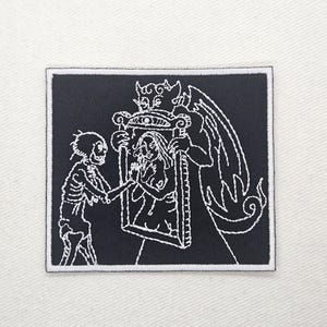 May include: Square black embroidered patch with a white border. The design features a skeleton reaching towards a woman in a frame held by a devil-like figure. The image is stitched in white thread on a black background.