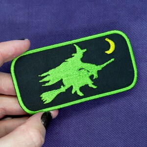 May include: A green embroidered patch of a witch flying on a broomstick with a crescent moon in the background. The patch is on a black background with a green border.