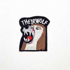 Op de afbeelding: Een geborduurde patch met een bruine en witte wolf met een open bek die scherpe tanden en een rode tong laat zien. De tekst THEIRWOLF is in witte letters op een zwarte achtergrond bovenaan de patch gestikt, die een zwarte rand heeft.