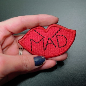 Op de afbeelding: Rode vilt patch in de vorm van lippen met het woord "MAD" geborduurd in zwart garen.