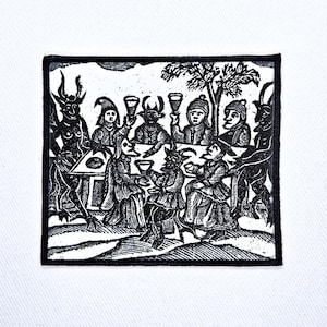 May include: A rectangular black and white embroidered patch featuring a detailed woodcut-style illustration of several figures, including horned creatures and people, gathered around a table with raised glasses in a toast under a tree.