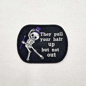 May include: Black embroidered patch featuring a skeleton wearing a top hat and holding a cane. The text reads "They pull your hair up but not out."