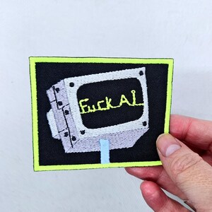 Puede incluir: Parche bordado con un diseño de pantalla de ordenador retro y el texto "Fuck AI" en amarillo. El parche tiene un fondo negro, un borde verde neón y un soporte azul claro.