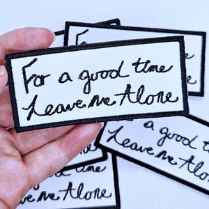 Può includere: Una toppa rettangolare ricamata con sfondo bianco e un bordo nero spesso. La toppa presenta un testo nero in corsivo che recita "For a good time leave me Alone" centrato sul tessuto. Il design è pulito, audace e ad alto contrasto.