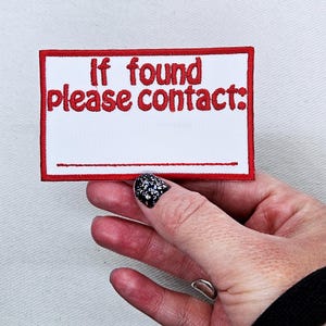 May include: Embroidered patch with a white background and a red border. The text "If found please contact:" is in red. A blank space is provided for contact information. The patch is held by a hand.
