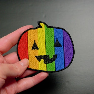 Może przedstawiać: Naszywka z haftem w kolorach tęczy przedstawiająca dynię z Halloween z czarnym, wyszytym uśmiechem.
