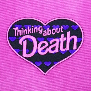 以下が含まれることがあります： 縁に紫色のハートが並んだ、黒いハート型のワッペン。ピンク色のアウトラインで縁取られています。「Thinking about Death」という文字がピンク色の文字で書かれています。