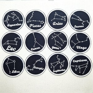 May include: A collection of twelve embroidered patches featuring zodiac constellations. Each patch is a black circle with a white border, displaying a constellation and its name in white script. The patches are arranged in a grid.
