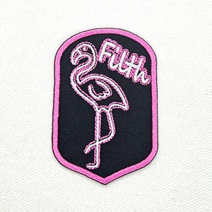 Op de afbeelding: Roze en witte geborduurde patch met een flamingo-silhouet en het woord "Filth" in een cursief schrift.