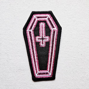 Op de afbeelding: Roze en witte geborduurde patch van een kist met een omgekeerd kruis erin. De patch is zwart met roze en witte stiksels.