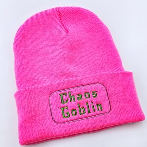 May include: A bright pink beanie hat with a folded brim. The hat features the embroidered text "Chaos Goblin" in green letters on a rectangular patch. The hat is made of a soft, knitted material.