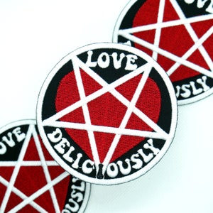 Puede incluir: Un parche bordado en blanco y negro con un corazón rojo dentro de un pentagrama. El texto "LOVE DELICIOUSLY" está escrito alrededor del pentagrama.