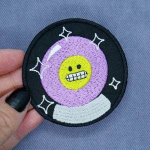 Könnte beinhalten: Ein gestickter Patch, der eine magische Acht-Kugel mit einem lächelnden Gesicht darin zeigt, in Lila und Gelb.