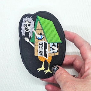 Puede incluir: Parche bordado con un diseño de casa caprichosa. La casa tiene un tejado verde, un ojo y una puerta en espiral, con patas de pollo. Una figura fantasmal está al lado de la casa. El parche está sobre un fondo ovalado negro.