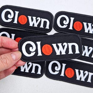 May include: A rectangular embroidered patch with a black background and white text that reads Clown, featuring a solid orange circle replacing the letter o. The patch has a black stitched border and is shown in a group of several identical items.