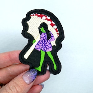 以下が含まれることがあります： 緑色の刺繍パッチ。長い黒髪の女性が白と赤の傘を持っています。女性は花柄の紫色のドレスを着ています。