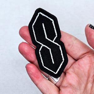 May include: Black patch featuring the stylized "S" design, outlined in white stitching. The patch has a black background and a white stitched border. The patch is held in a hand.