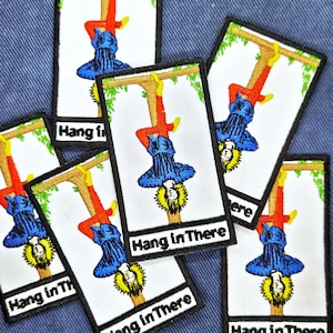 May include: Rectangular white patches with a black border, featuring a tarot card design. The image depicts a figure hanging upside down, with the text "Hang in There" below. The figure is wearing blue and red clothing.