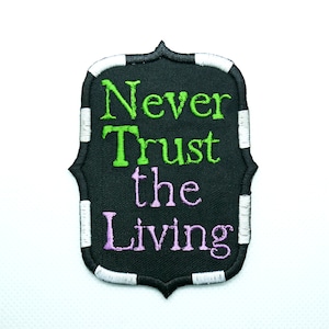 Może przedstawiać: Czarno-biała naszywka haftowana z tekstem "Never Trust the Living" w zielonych i fioletowych literach.