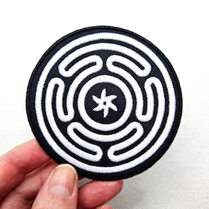 Può includere: Toppa ricamata in bianco e nero con un design circolare. Il centro del cerchio presenta una stella a cinque punte. L'anello esterno del cerchio presenta una serie di cerchi concentrici.